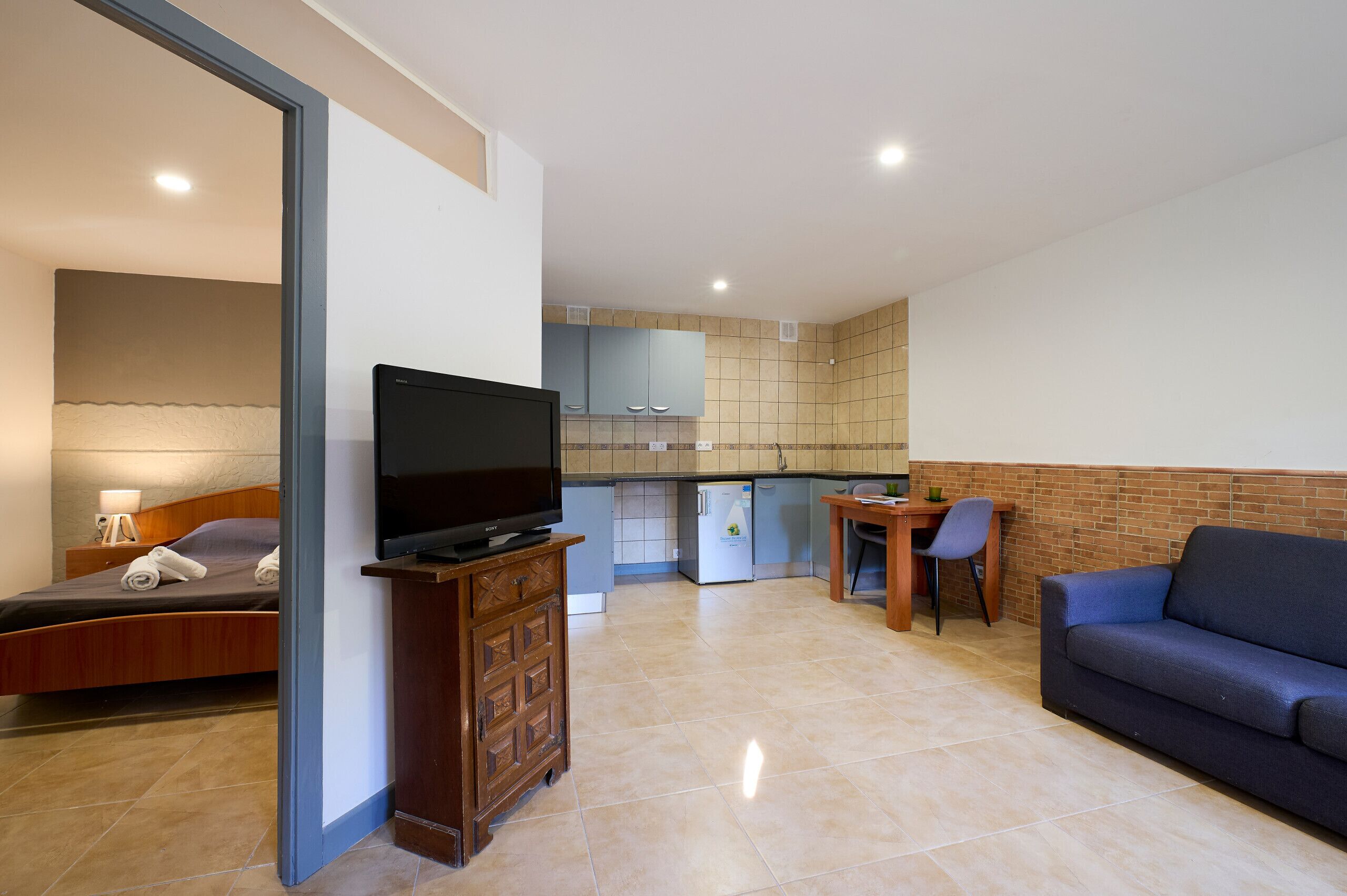 A modern living room with a sofa, a television on a wooden cabinet, a bed with a nightstand, a small kitchen area with cabinets and a refrigerator, and a dining table with a chair.
