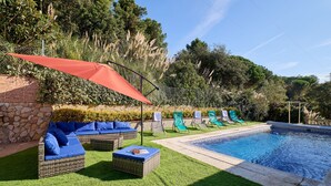 Ein Poolbereich mit einer Backsteinmauer, einem roten Sonnenschirm, Korbmöbeln und grünen Liegestühlen.