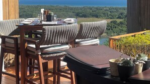 Un terrazzo in legno con mobili in vimini e un tavolo apparecchiato per la cena, con vista su una città costiera e montagne.