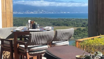 A wooden deck with wicker furniture and a table set for dining, overlooking a coastal city and mountains.