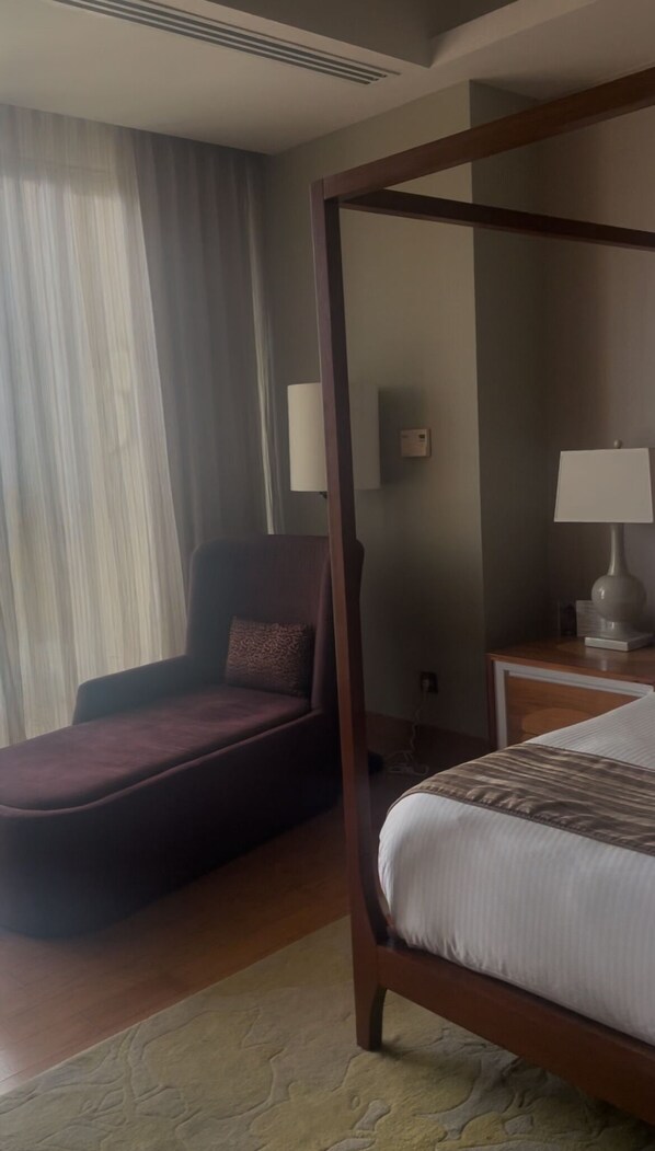 A bedroom with a four-poster bed, a chaise lounge, a bedside table with a lamp, and a window with curtains.
