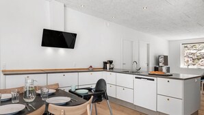 Une cuisine moderne avec des meubles blancs, un téléviseur à écran plat fixé au-dessus du comptoir, et un coin repas avec une table et des chaises en bois.