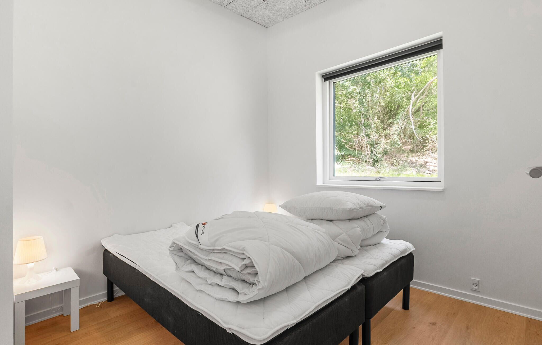 A minimalist bedroom with a bed, a bedside table with a lamp, and a window with a view of greenery.