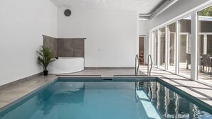 Une piscine intérieure moderne dotée d’un jacuzzi, de portes en verre transparent donnant sur un espace extérieur et d’un plafond équipé d’un éclairage encastré.