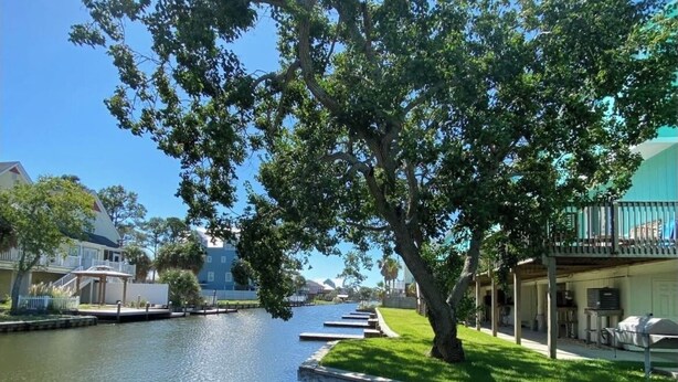 A riverside view with a dock, grassy areas, and buildings in the background. - Charming 2bedroom condo on the canal a dock available. In beautiful Gulf Shores! (Gulf Shores)