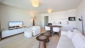 A modern living room with a flat-screen TV, a sofa, two round wooden tables, and a kitchen area with white cabinets and a microwave. - Lovely apartment in Oostende (Oostende)