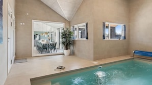 Un espace piscine intérieur moderne, offrant une vue dégagée sur le bassin, un coin repas avec une table et des chaises, et une grande plante en pot.