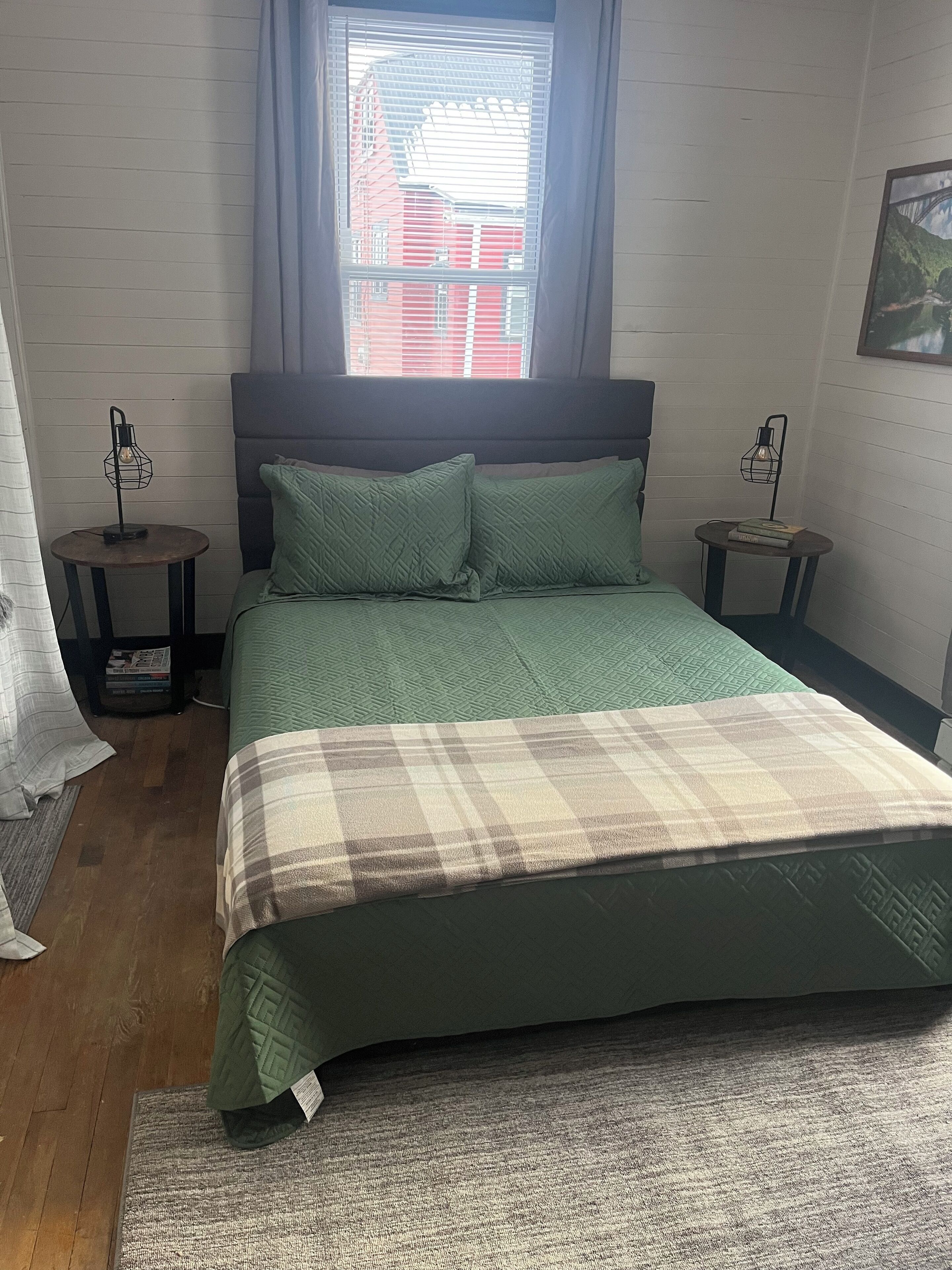 Ein Schlafzimmer mit einem Bett, zwei Nachttischen, einem Fenster mit Jalousien und Blick auf Gebäude.