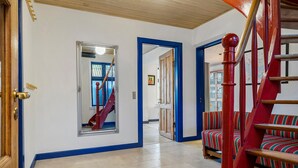Un couloir avec une porte en bois, un miroir, un escalier avec des rampes rouges et un banc à rayures.