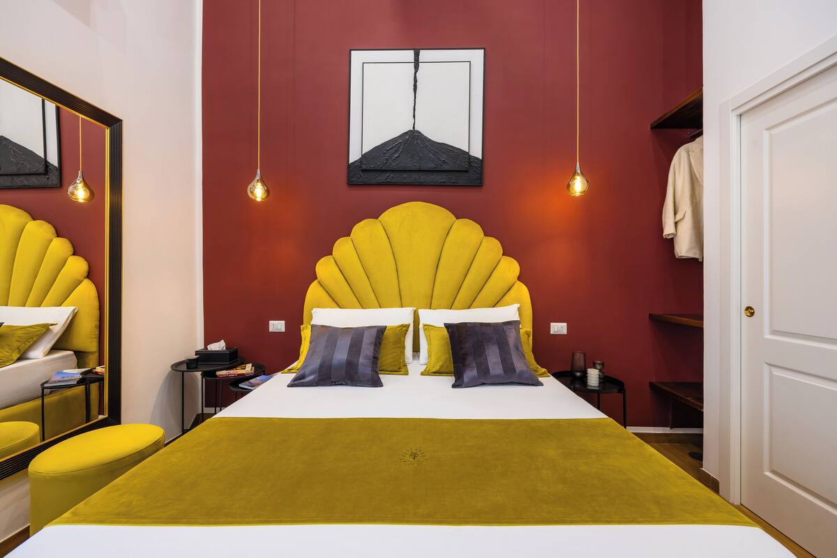 A hotel room with a large bed, yellow headboard, and a red accent wall.