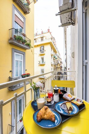 A yellow balcony with breakfast items, including croissants, a cake, and a coffee drink, overlooking a narrow street with yellow buildings. - L Angolo do core (Naples)