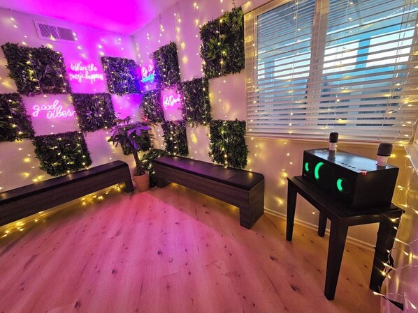 Una habitación con piso de madera, un banco, una mesa con un altavoz y paredes decoradas con plantas y luces de cuerda.