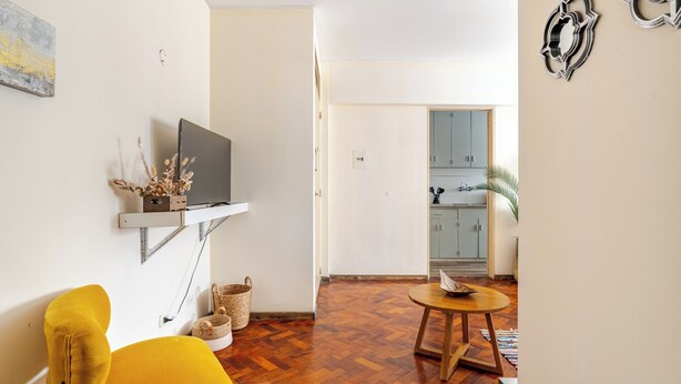 Ein Wohnzimmer mit Holzboden, einem gelben Sessel, einem Fernseher an einer Wandhalterung und einem kleinen runden Tisch mit Tablett.
