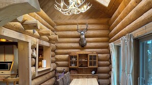 Ein rustikales Blockhaus-Interieur mit Esstisch, Holzbänken, einer Kristallleuchtre, einem aufgehängten Hirschgeweih und einem großen Fenster.