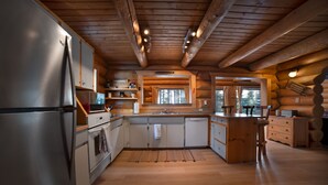 Eine Holzblockhausküche mit Holzbalken, einem Edelstahlkühlschrank, Holzausstattung und einem großen Fenster.