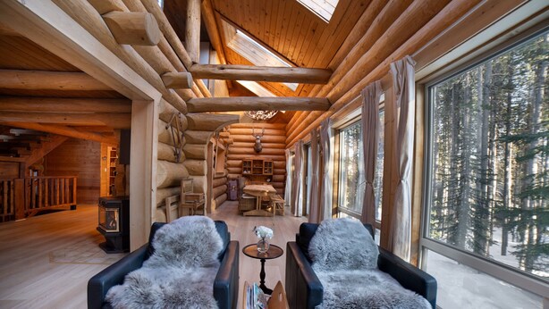 Ein Holzblockhaus-Interieur mit einer gemütlichen Sitzecke, einem Kamin und einem großen Fenster mit Blick auf den Wald.