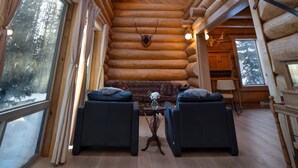 Ein rustikales Blockhaus-Interieur mit hohen Decken, Holzbalken, zwei schwarzen Sofas, einem kleinen Tisch mit einer Vase und großen Fenstern mit Blick auf verschneite Landschaft.