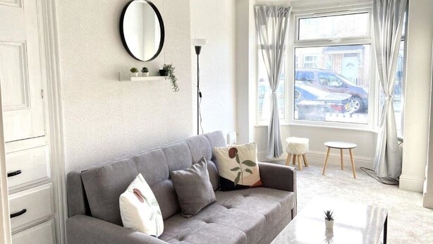 A modern living room with a grey sofa, a round mirror, a small plant shelf, and a view of the street through a window with curtains. - Kisobi Home Bedroom 1 (Kingston upon Hull)