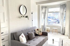 Un salon moderne avec un canapé gris, un miroir rond, une petite étagère pour plantes et une vue sur la rue à travers une fenêtre avec des rideaux.