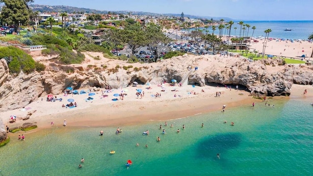 A coastal beach with clear turquoise waters, rocky cliffs, and a view of a residential area in the background. - Cozy, private & charming 3bd/2ba cottage in a magical little village by the Sea (Newport Beach)