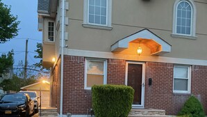 Une maison à deux étages avec une façade en briques, une porte d’entrée et un petit jardin devant, avec une pelouse bien entretenue et un arbuste taillé.