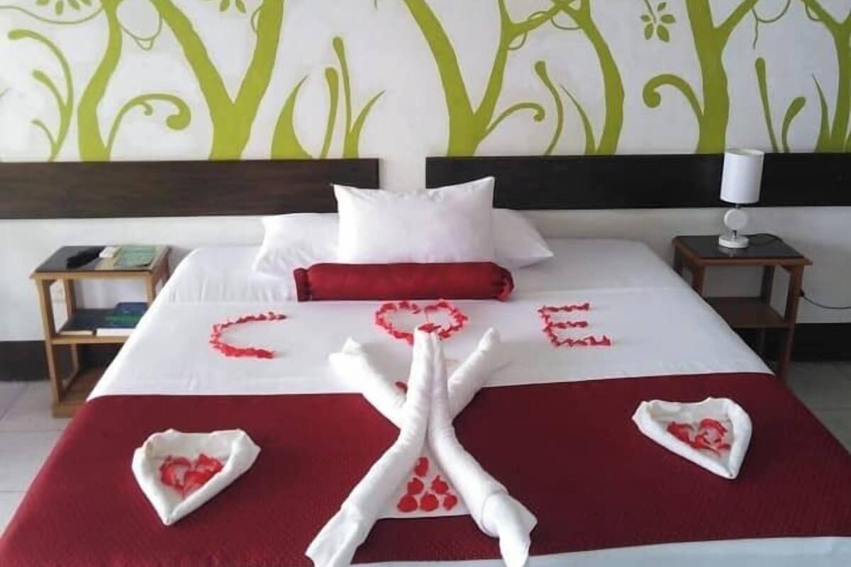 Una cama bien hecha con una colcha rojo y blanco, almohadas blancas y un arreglo de pétalos de rosa roja en forma de corazón sobre la cama.