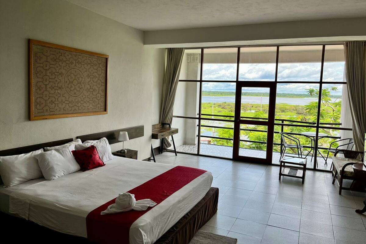 Un dormitorio espacioso con una cama grande, un escritorio y vistas a un paisaje exuberante a través de amplios ventanales.