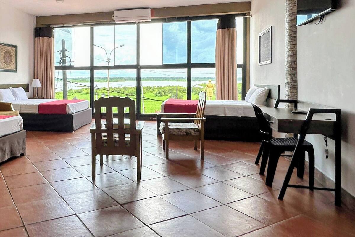 Habitación de hotel con dos camas, una silla de madera, un escritorio con silla y ventanas grandes que dan a zonas verdes.
