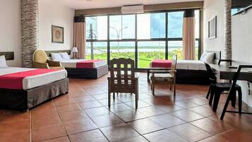 Habitación de hotel con dos camas, una silla de madera, un escritorio con silla y ventanas grandes que dan a zonas verdes.