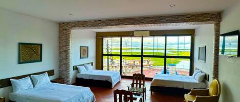 Habitación de hotel con dos camas, una silla de madera y vistas a un paisaje verde a través de amplios ventanales.
