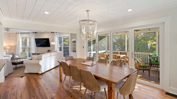 A spacious dining area with a wooden table and wicker chairs, a large chandelier, and a view of the outdoors through large windows.