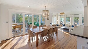 A spacious dining area with a large wooden table and wicker chairs, a chandelier, and a view of a deck and greenery outside.