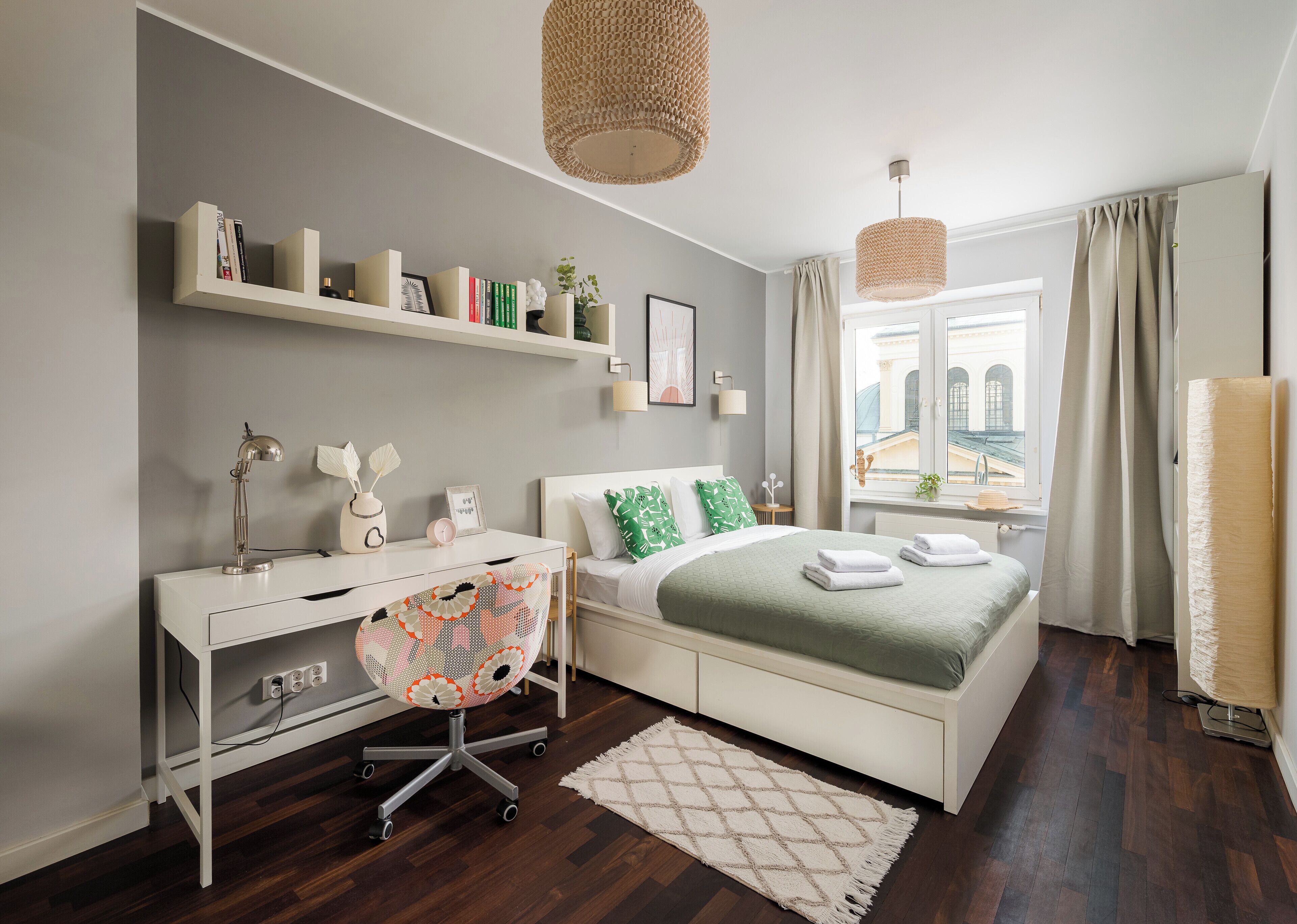 Une chambre à coucher moderne avec un lit blanc, un bureau et une chaise. On y trouve des étagères remplies de livres et d’objets décoratifs, une fenêtre avec vue et un luminaire suspendu.