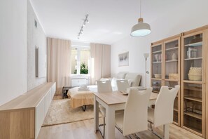 A modern hotel room with a dining area, a sofa, a cabinet with glass doors, and a window with curtains. - Spodek 2-Bedroom Apartment (Katowice)