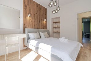 A modern bedroom with a wooden accent wall, a grey sofa bed, a white chair, a bedside table, and a hanging light fixture. - Spodek 2-Bedroom Apartment (Katowice)
