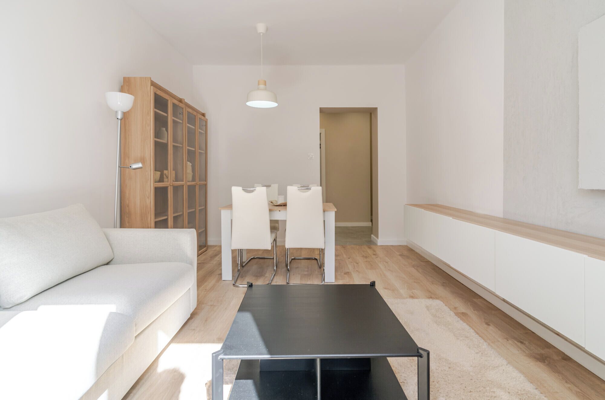 A modern living room with a white sofa, a dining table with chairs, a black coffee table, and a wooden cabinet with glass doors.