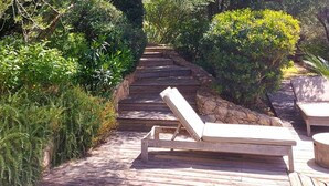 Una terraza de madera con una silla de descanso y una mesa, rodeada de exuberante vegetación y un muro de piedra.