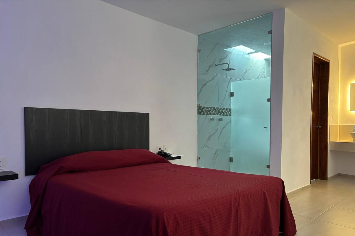 Un dormitorio con cama, mesita de noche, cabina de ducha de vidrio y puerta de madera.