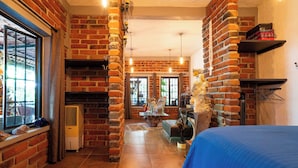 A room with exposed brick walls, a fireplace, a bed with a blue bedspread, and a window with a view of the outdoors. - Cozy House with Pool and 3 Bedrooms (Yautepec)