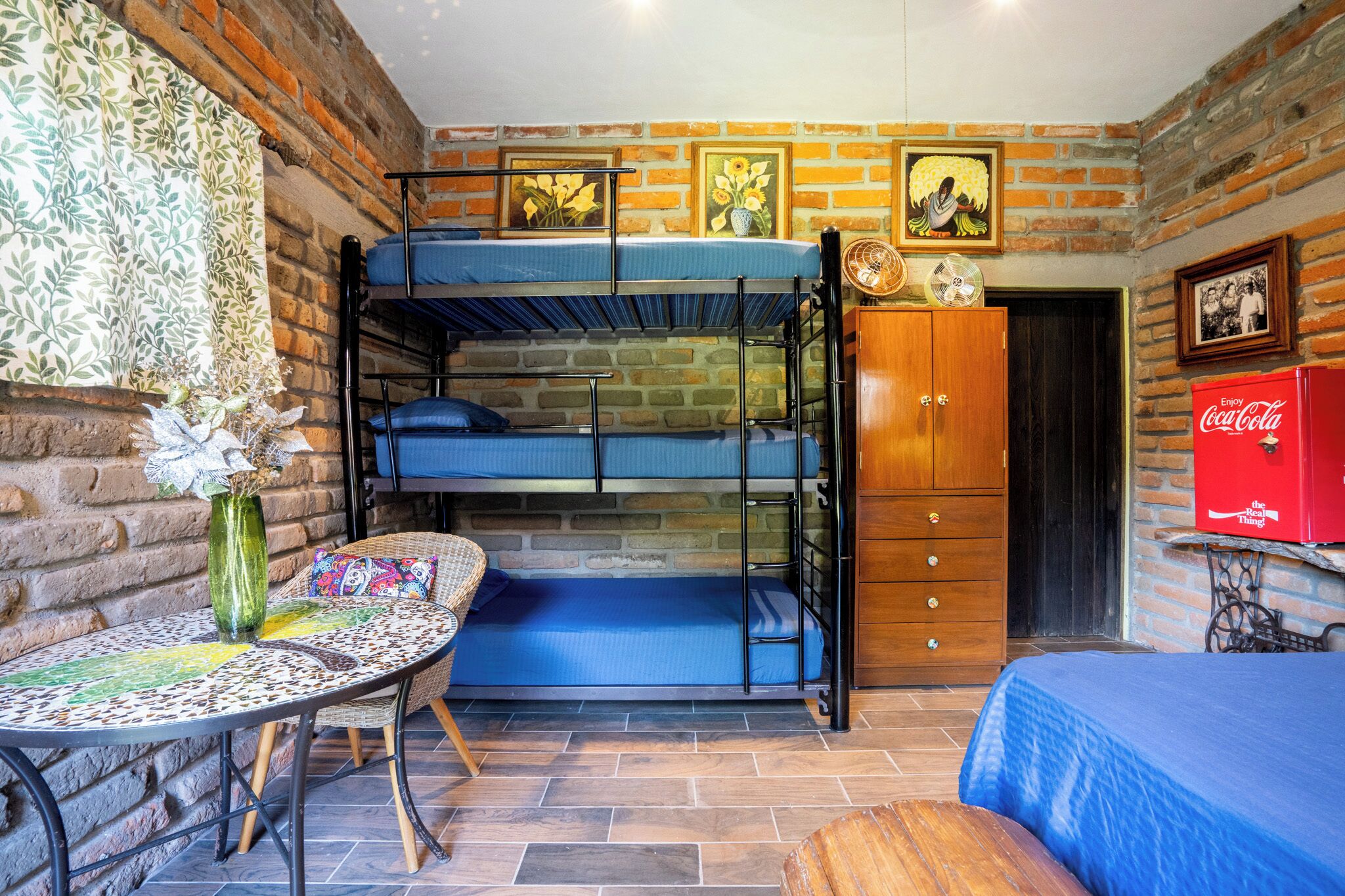 Una habitación con una cama litera, un armario de madera, una mesa con mantel estampado y un refrigerador rojo Coca-Cola.