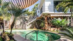 A pool area with a stone wall, palm trees, and a staircase leading to a balcony. - Cozy House with Pool and 3 Bedrooms (Yautepec)