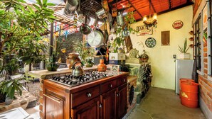 A kitchen with a gas stove, hanging pots, and various kitchen utensils. There are potted plants and a refrigerator in the background. - Cozy House with Pool and 3 Bedrooms (Yautepec)