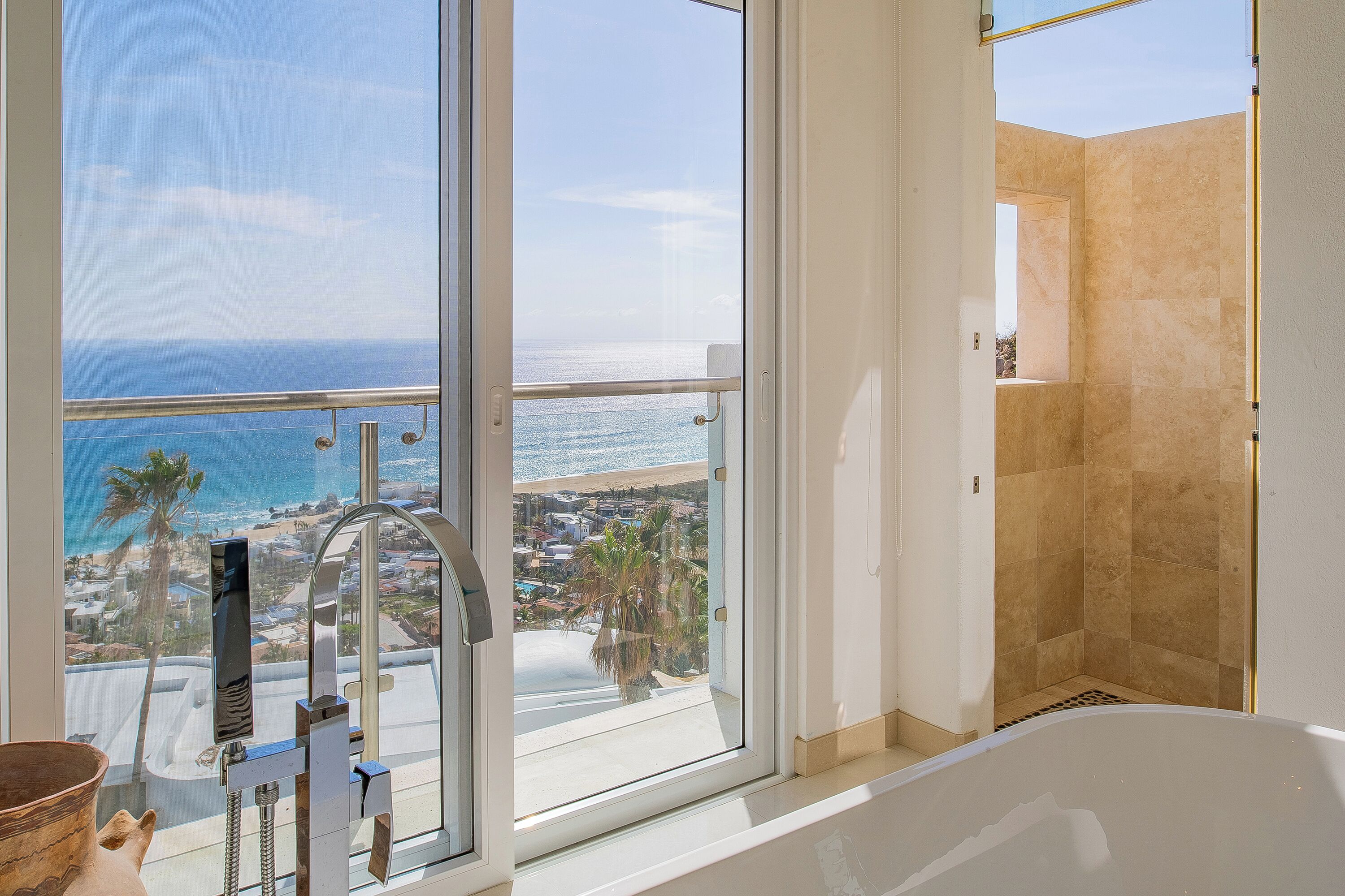 A bathroom with a bathtub, a shower, and a large window offering a view of the ocean and a coastal town.