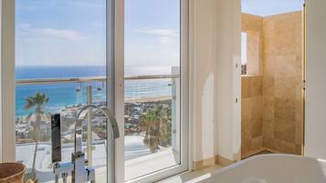 A bathroom with a bathtub, a shower, and a large window offering a view of the ocean and a coastal town.