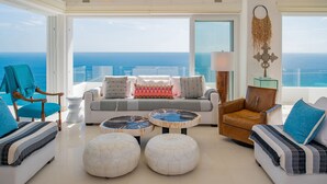 Een ruime woonkamer met een groot raam met uitzicht op de oceaan, ingericht met een bank, fauteuils, poefs en een salontafel.