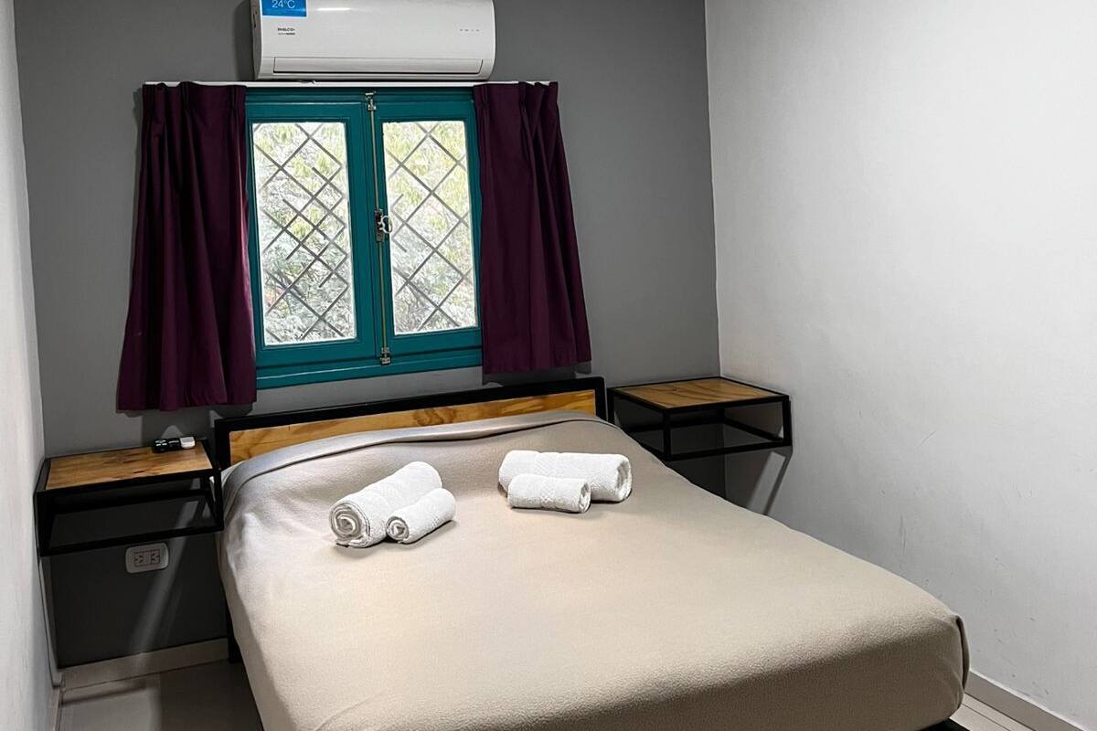 A single bed with a headboard, a window with curtains, a wall-mounted air conditioner, and a bedside table with a lamp.
