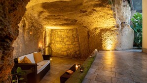 Tunnelmallinen, luolanomainen sisustus, jossa on kiviseinä, rottinkisohva tyynyineen, pieni pöytä ja kynttilöitä.