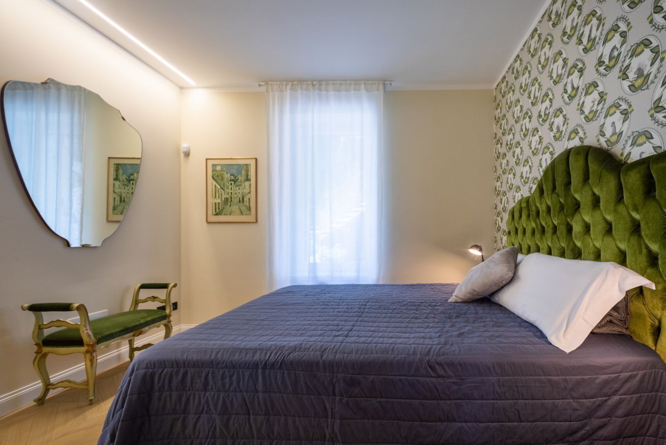 A bedroom with a large bed, a green upholstered headboard, a green chair, a mirror, and a window with sheer curtains.