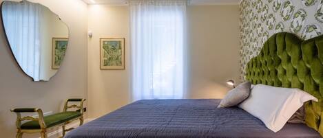 A bedroom with a large bed, a green upholstered headboard, a green chair, a mirror, and a window with sheer curtains.