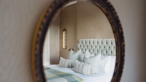 Une chambre à coucher avec une tête de lit capitonnée, du linge de lit blanc et un miroir qui reflète la pièce.